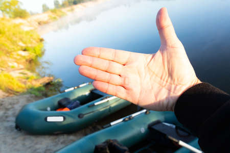The palm of a man's hand with a mazole from an oar against the background of two rubber inflatable boats with fishing tackle in the early morning, parked on the bank of the river.の写真素材