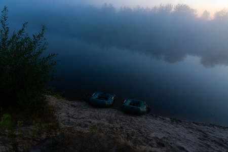 Two rubber boats with fishing tackle in the early morning during the fog, parked on the banks of the river.の写真素材