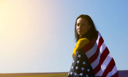 A young, dark-haired woman holding the flag of the United States of America against the sunny sky. Memorial Day and USA Independence Day concept.の写真素材