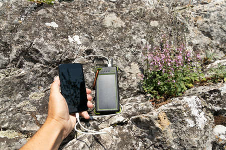 The smartphone is charged with a portable solar power bank charger on a rock during extreme travel.