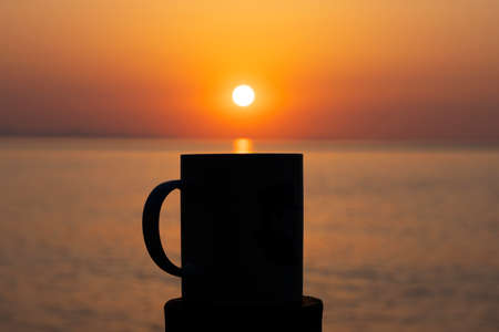 Selective focus image of a cup of coffee or tea at dawn in the morning over the sea and the rising...