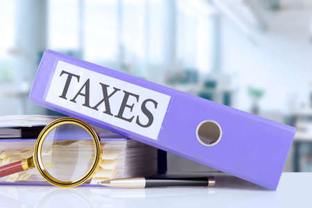Taxes, text written in a folder with documents in trendy purple color, lying on a stack of papers, documents on an office desk and a magnifying glass, against a soft blur office background. high quality photoの写真素材