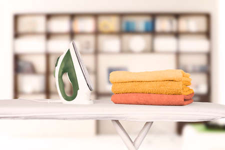 An electric iron stands on an ironing board and folded bath towels in orange and shades against the background of a home interior, office, library. Blurred background, soft focus. high quality photoの写真素材