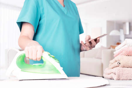 Caucasian woman, housewife, housekeeper, maid ironing clothes with an electric iron on an ironing board and looking at the screen of a mobile phone. home interior background. soft focus. high quality photoの写真素材