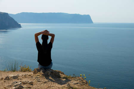 A beautiful woman with dark hair and hands in her hair sits on the edge of a mountain against the...
