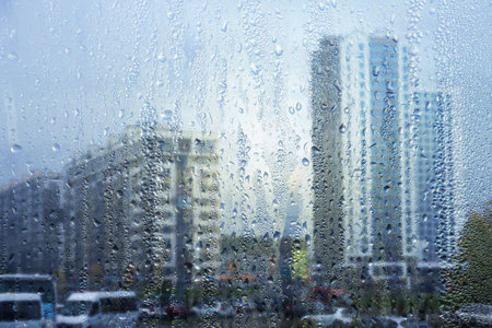 Rainy window, raindrops on the glass, against the background of a city street with houses and a road. Soft blurred background.の写真素材