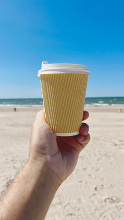 Brown paper cup with coffee in hand on the background of sandy beach and sea. Baltic Sea