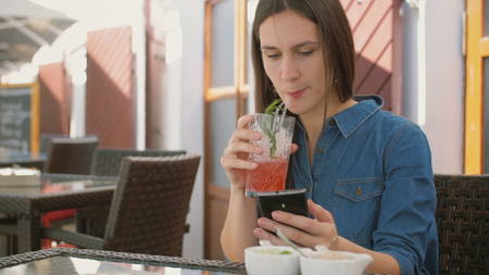 Brunette girl in a dress uses smart phone while sitting outside in a cafe, drinking and enjoying a cool drink from a straw tube hot summer day. 4k,の写真素材