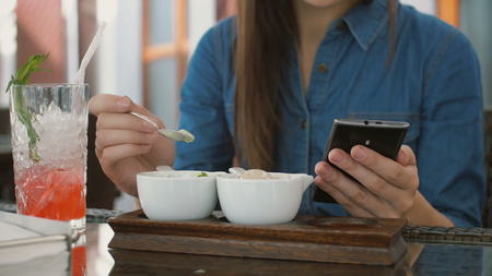 Brunette girl in a dress uses smart phone while sitting outside in a cafe, eating ice cream. 4k,の写真素材