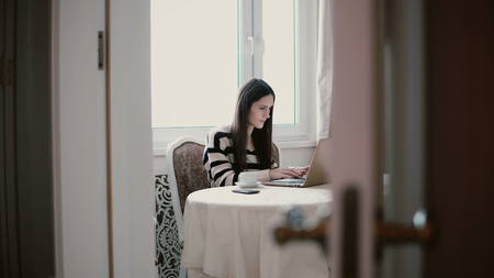 Attractive young woman uses laptop and enjoying morning coffee on a beautiful bright kitchen. view through the open doorsの写真素材