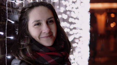 portrait of a young attractive woman in the falling snow during Christmas night looking at the camera standing near colorful sparkling lights wallの写真素材