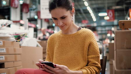 young attractive woman in the orange jacket uses a smartphone, sitting among the things in a store or warehouseの写真素材