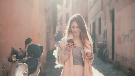 Young happy woman walking in city centre at morning, using smartphone. Girl surfing the Internet, texting with someone.の写真素材