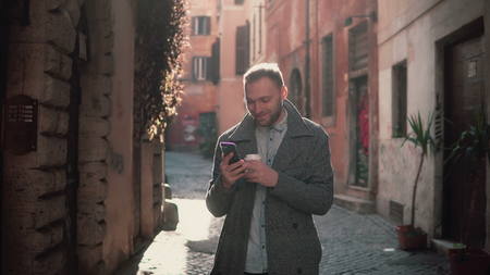 Young smiling man walking in city at morning and using smartphone. Guy browse the Internet, texting with someone.の写真素材