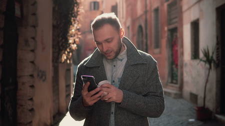 Young attractive man walking in city and using smartphone. Male chatting with friends and drinking coffee.の写真素材