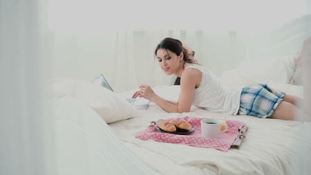 Young woman using laptop during breakfast lying on white bed at home. Happy girl eat croissant and browses the Internet.の写真素材