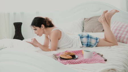 Beautiful girl lying on the bed in morning and chatting, using laptop for that. Young woman eating croissant and typing.の写真素材