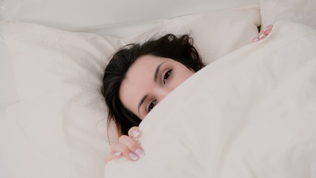 Close-up of a young beautiful woman with a funny look hiding under the blanket. Girl having fun in bed.の写真素材