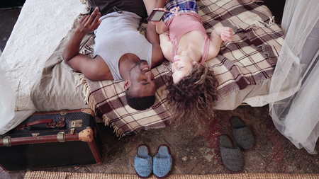 Multiracial couple lying on bed, listening music on smartphone. Man and woman in pajamas spending...