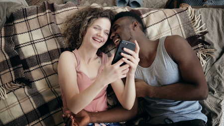 Multiethnic couple lying on bed and using smartphone. Man and woman happy together. Male and female laughing, smiling.の写真素材