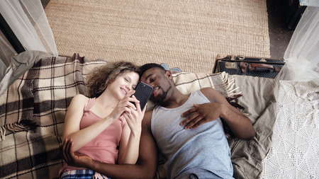 Multiracial couple lying on bed. Man and woman spend time together with joy and fan, use Smartphone, surf the internet.の写真素材