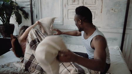 Multiracial couple spending time together. Male and female lying on bed, laughing. Man and woman fighting with pillows.の写真素材