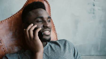 Portrait of young African male sitting in the chair and talking on Smartphone. Man laughs and nods his head agreeing.の写真素材