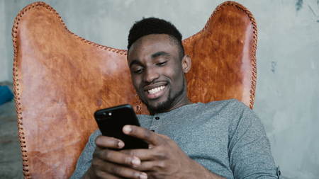Portrait of African man sitting in chair, using Smartphone. Man reads messages and laughs. Guy typing massages, smilingの写真素材