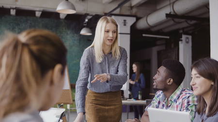 Young blonde woman manager talking with mixed race team. Businesswoman giving direction to colleagues at modern office.の写真素材