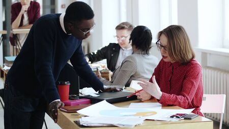 Happy young black male manager talking to middle aged European boss woman, multiethnic people at modern healthy office. Casual African man working together with beautiful female leader in red dress.の写真素材