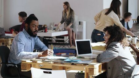 Happy young positive African man smiling, talking to beautiful Caucasian female manager at trendy loft office table.の写真素材