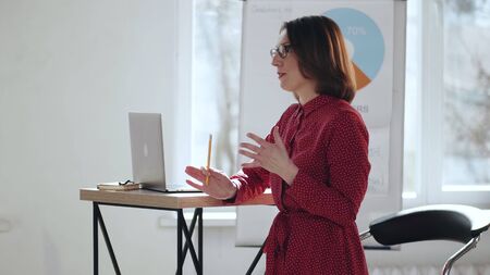 Happy beautiful middle aged Caucasian coach business woman in eyeglasses giving seminar talk at light modern office.の写真素材