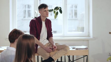 Happy young start-up company employee sitting on table at modern loft office listening to seminar talking to colleagues.の写真素材