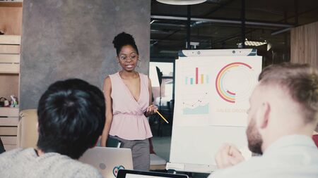 Female team leader explaining data on flip chart. Modern multiethnic financial business meeting in trendy coworking 4K.の写真素材