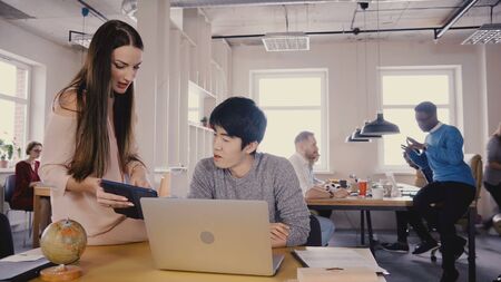 Female boss gives instructions to Asian man. Multiethnic colleagues work in casual modern loft...