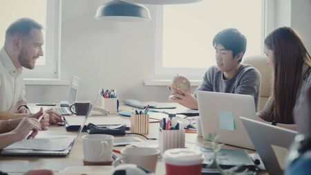 Happy Asian businessman takes a little globe, points at country at multiethnic office meeting in...