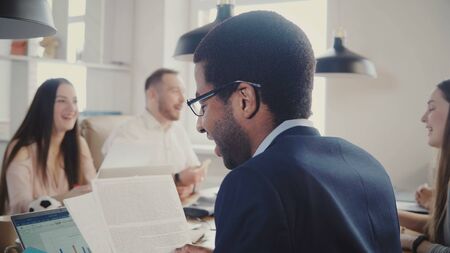Back view African American businessman reading through documents at multiethnic office meeting, diagrams on laptop 4K.の写真素材