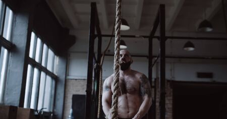 Shirtless young athletic Caucasian sportsman climbing rope during extreme functional physical workout in large gym.の写真素材