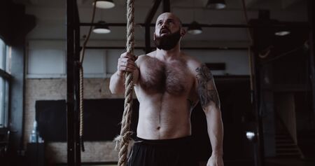 Shirtless young athletic Caucasian sportsman climbing rope during extreme functional physical workout in large gym.の写真素材