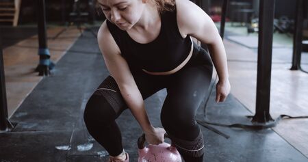 Young athletic red haired professional athlete woman lifting kettlebell weight in large atmospheric gym slow motion.の写真素材