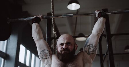 Close-up young Caucasian athlete man lifting heavy barbell in large hardcore gym hall. Ambition and...