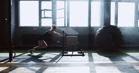 Sliding side view young athletic Caucasian woman exercising with heavy training sled in big...