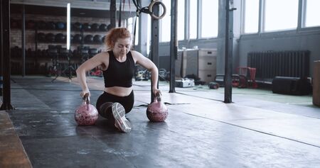 Young professional athletic red haired coach woman doing split stretch exercise on kettlebells in large gym slow motion.の写真素材