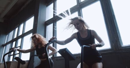 Two young athletic Caucasian women doing cardio workout on gym bike machines with fans slow motion....