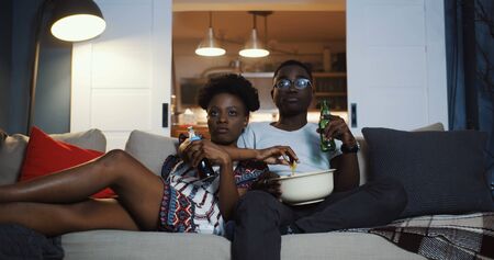 Happy young African positive friends couple watching TV at home on the couch, eating snacks and laughing slow motion.の写真素材