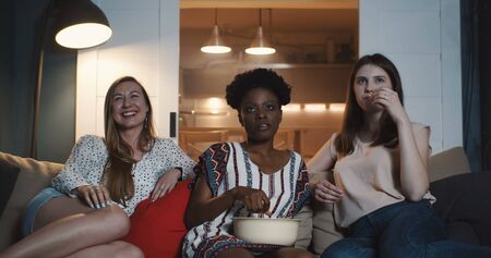 Three happy young multiethnic women express different emotion watching movies at home with snacks slow motion.の写真素材