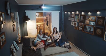 Beautiful happy multiethnic women friends laugh watching comedy TV show at home on big screen with projector slow motionの写真素材