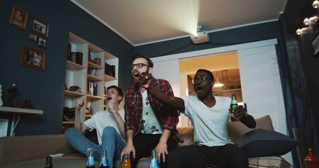 Happy multiethnic male friends shout and cheer watching sports game together at home on TV using projector slow motion.の写真素材