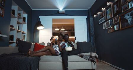 Happy young relaxed black couple of friends sit together on couch at home, watch TV with drinks and popcorn slow motion.の写真素材