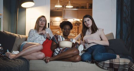 Sad beautiful multiethnic women friends wipe tears with tissue, cry watching emotional drama movie at home slow motion. Young female students enjoying favourite moments of romantic film.の写真素材